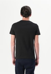 Man med kort mörkt hår klädd i en enkel svart t-shirt och marinblå byxor, stående vänd bort från kameran mot en vit bakgrund.