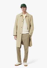 Beige trench coat over en hvit genser i strikk, lyse grønne detaljer, sammen med beige bukser og hvite joggesko med brune detaljer; grønn caps.