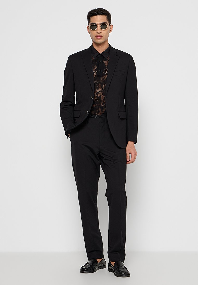 Un uomo è in piedi in un completo nero su misura con una camicia a fantasia sotto, indossando occhiali da sole e scarpe eleganti, su uno sfondo bianco uniforme.