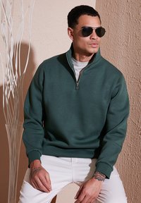 Felpa verde con zip e finitura texturizzata, indossata sopra una maglietta grigia chiara, abbinata a pantaloni bianchi.