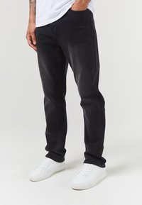 Los jeans de mezclilla negros presentan un corte ajustado con una sutil textura desgastada, acompañados de zapatillas blancas para un look informal.