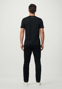 Mies seisoo selkä kameralle, päällään musta T-paita, jossa on kauluksessa teksti "HACKETT", mustat farkut ja valkoiset lenkkarit yksinkertaista taustaa vasten.