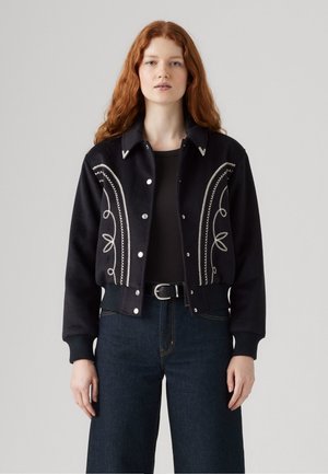 Zwarte cropped jacket met zilveren geborduurde patronen en witte accenten. Heeft een knoopsluiting en ribgebreide manchetten. Gecombineerd met hoge taille jeans.