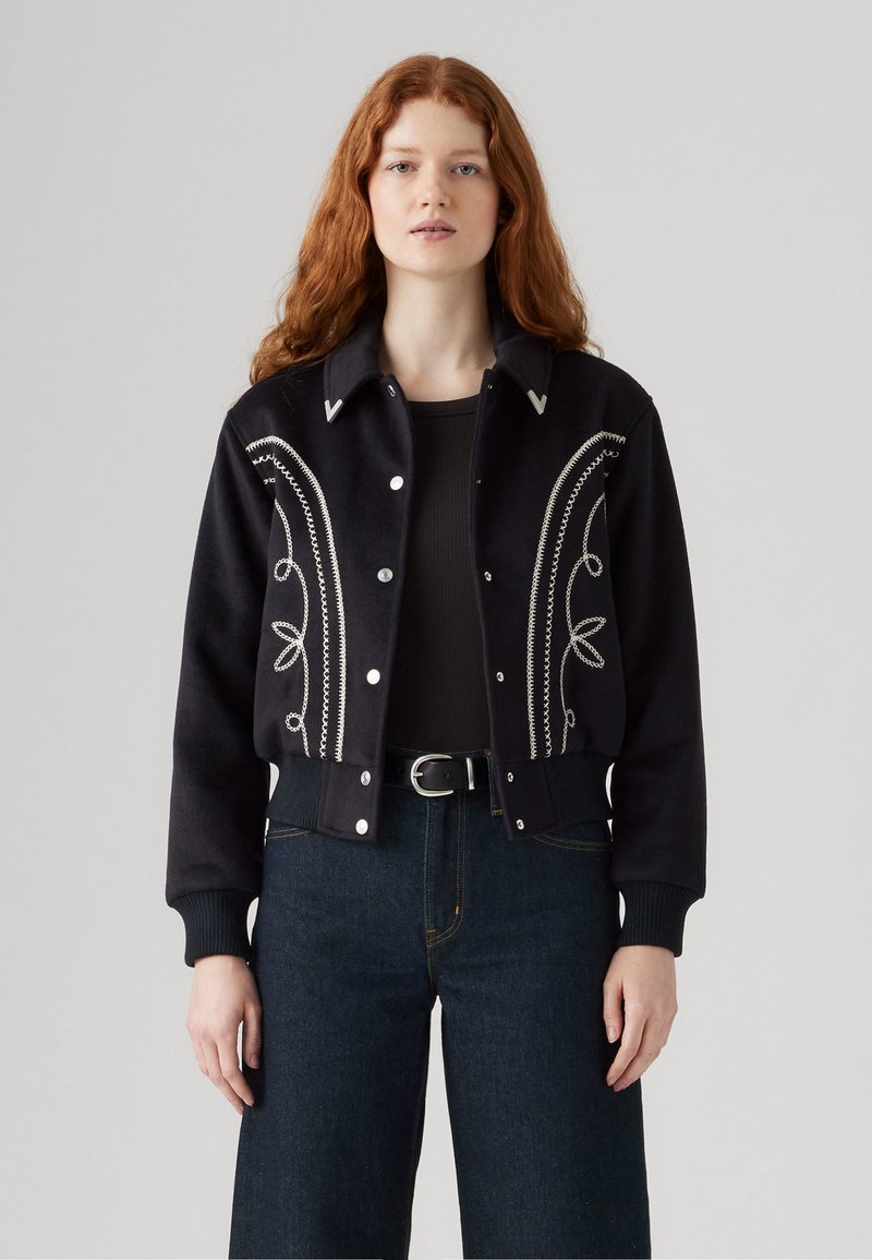 Veste noire courte avec des motifs brodés en argent et des accents blancs. Comprend une fermeture à boutons et des poignets côtelés. Associée à un jean taille haute.