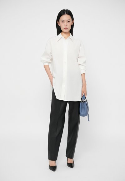 Biela košeľa s gombíkmi a vyhrnutými rukávmi, kombinovaná s voľnými čiernymi nohavicami a špicatými čiernymi podpätkami. V jednej ruke drží malú modrú kabelku.