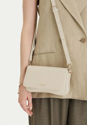 Béžová kožená kabelka přes rameno s klopou a zlatým logem LASOCKI, nošená s béžovým blejzrem bez rukávů a hnědými kalhotami se vzorem rybí kost.