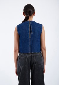 Top de mezclilla recortado; lavado oscuro, diseño sin mangas; cuenta con una cremallera central en la parte posterior con detalles de dobladillo sin terminar; combinado con jeans negros de cintura alta.