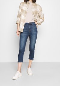 Camisa de cuadros beige con botones sobre una blusa beige clara, combinada con jeans cortos de tiro alto de color azul oscuro y deportivas blancas.
