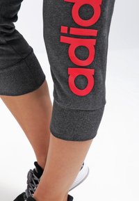 Grijze sportieve cropped broek met ribbels aan de boorden en een groot rood "adidas" logo dat verticaal langs de zijkant loopt. Zwarte sneakers met een gestructureerd ontwerp.