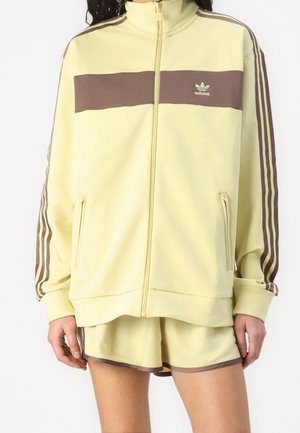 Kvinde iført en lysegul Adidas lynlåstrøje med brune striber og matchende shorts med brun kant.