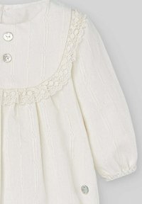 Biała bawełniana sukienka z koronkową lamówką, plisami, okrągłym kołnierzykiem, trzema guzikami i marszczonymi rękawami. Z ozdobnym guzikiem przy dolnym brzegu.