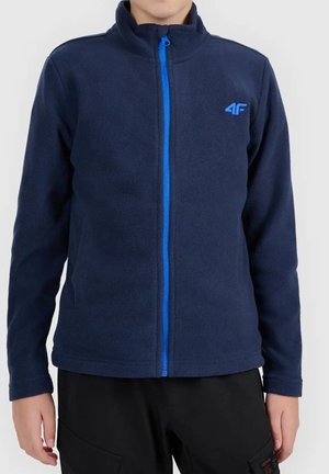 Enfant portant une veste polaire bleu marine avec une fermeture éclair bleu vif et un logo 4F sur la poitrine gauche, associée à un pantalon noir.