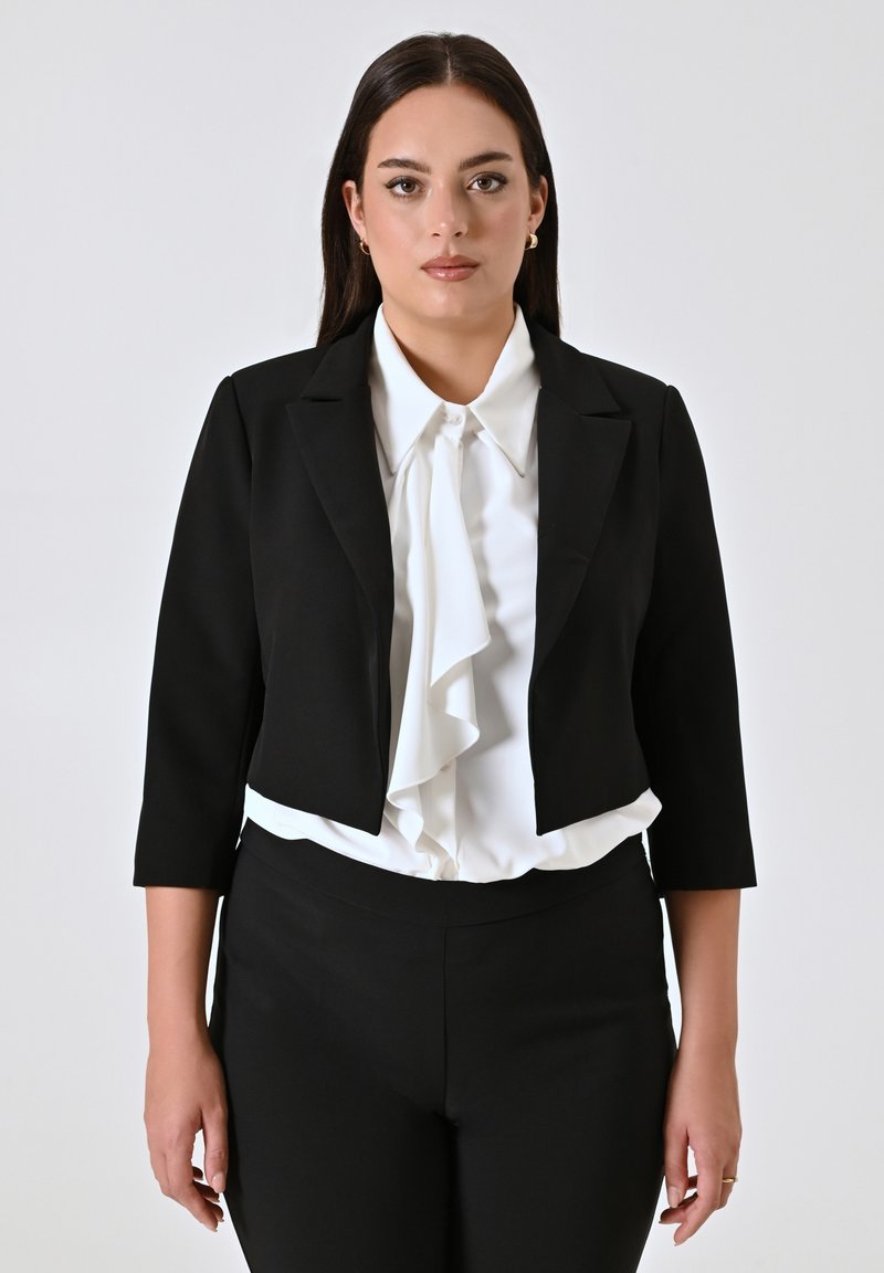 Zwarte cropped blazer over een witte gerimpelde blouse, gecombineerd met zwarte broeken. De blazer heeft driekwart mouwen en een klassieke kraag.