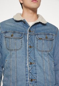 Gai zilas džinsu jaka ar pūkainu baltu apkakli, priekšējiem kabatām un brūnajiem pogām. Iezīmēta redzamā šuvuma un tekstūras auduma.