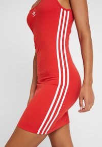 Kobieta w dopasowanej czerwonej sukience Adidas z trzema białymi paskami po bokach, stojąca z jednym ramieniem swobodnie opuszczonym wzdłuż ciała.
