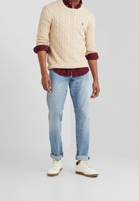 Miesten kermaisen värinen kylkineulepusero viininpunaisen paidan päällä, yhdistetty vaaleansiniin farkkuihin ja valkoisiin lenkkareihin, joissa on ruskeat pohjat.