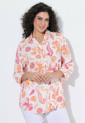 Kvinde med krøllet mørkt hår iført hvid skjorte med knapper med pink og orange mønstre af havdyr og skaller, stående mod en hvid baggrund.