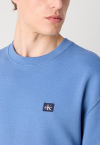 Blue sweatshirt featuring a round neckline and ribbed cuffs. Includes a small, dark blue Calvin Klein logo patch on the chest.