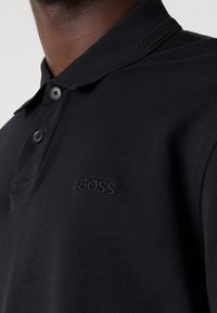 Polo noir avec col côtelé et deux boutons, logo brodé "BOSS" sur la poitrine, porté par une personne à peau foncée.