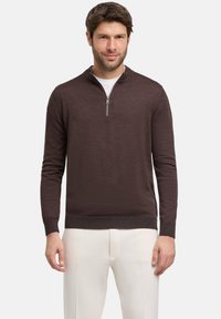 Maglione zip-neck marrone scuro realizzato in tessuto testurizzato, con maniche lunghe e orlo aderente, indossato sopra una maglietta bianca e pantaloni chiari.