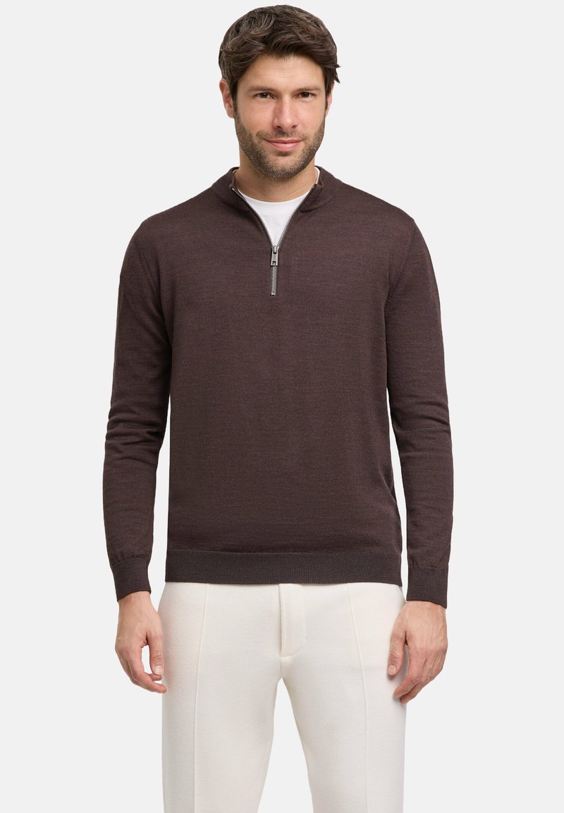 Maglione zip-neck marrone scuro realizzato in tessuto testurizzato, con maniche lunghe e orlo aderente, indossato sopra una maglietta bianca e pantaloni chiari.