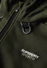 Felpa con zip di colore verde scuro realizzata in tessuto morbido, dotata di cappuccio con cordino e di un logo bianco che reca la scritta "SUPERDRY SPORT" sul davanti.