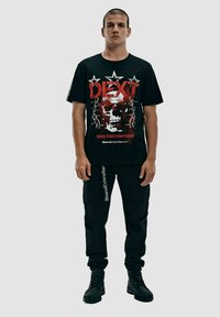 Czarna koszulka z grafiką z czerwonym tekstem i ilustracją czaszki, w połączeniu z czarnymi spodniami cargo i butami wojskowymi. Prosty, prosty krój.