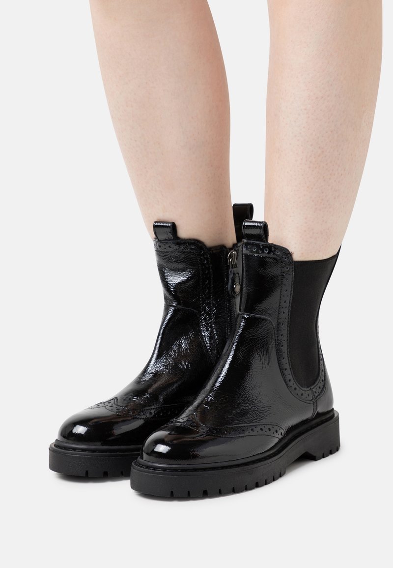 Bottines en cuir verni noir avec un motif brogue, des panneaux élastiques sur les côtés, et une semelle noire épaisse antidérapante.