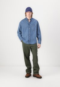 Cămașă din denim cu nasturi, albastră deschisă; pantaloni verzi; cizme maro cu șireturi; căciulă albastră cu nervuri. Modelul stă în fața unui fundal alb.