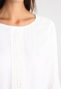 Blusa bianca con delicati dettagli ricamati lungo il centro e le maniche, realizzata in un tessuto morbido e liscio, con scollatura rotonda.