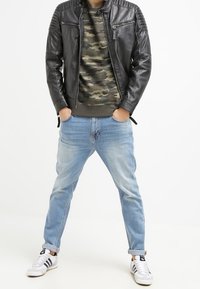 Chaqueta de cuero negra con acentos acolchados en los hombros, sudadera de camuflaje, jeans azules descoloridos y zapatillas blancas con rayas negras.