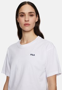 T-shirt bianco in cotone a maniche corte, con il logo FILA blu navy nella parte superiore sinistra del petto. Design classico con colletto a girocollo.