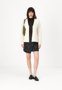 Cardigan a coste crema con bottoni, top aderente nero, pantaloni corti neri con pattern, calzini beige e mocassini neri. Borsa a tracolla verde.