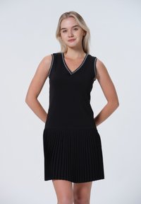 Robe noire sans manches avec un décolleté en V, présentant un buste ajusté et une jupe plissée. Bordée d'une couleur contrastante le long de l'encolure.
