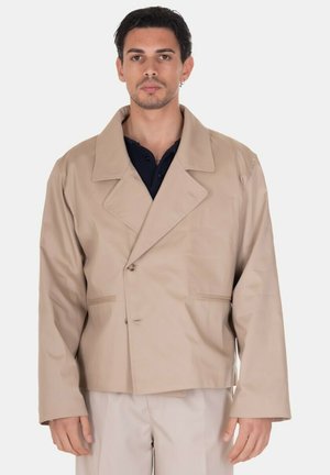 Uomo con blazer beige oversize sopra camicia nera, in piedi davanti a uno sfondo bianco semplice, che guarda dritto davanti a sé.