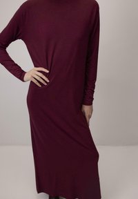 Robe à manches longues couleur bordeaux en tissu doux. Corsage ajusté se transformant en une jupe fluide et ample. Texture lisse avec un design simple.