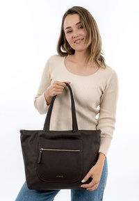 Tumši brūna ādas tote soma ar fakturētu virsmu, lielizmēra priekšējo kabatu, zelta rāvējslēdzēja detaļu un diviem izturīgiem rokturiem.