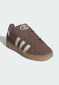 Brown suede sneaker with three white stripes, round logo on tongue, textured laces, and a gum rubber sole. Casual low-top design.