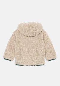 Giacca in pile beige e soffice con cappuccio, due cuciture laterali e polsini elastici verdi. Texture morbida con un taglio arrotondato e casual.