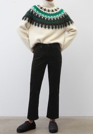 Une personne porte un pull à col roulé blanc avec des motifs verts et noirs, un pantalon noir et des chaussures noires à enfiler, debout avec les mains sur les hanches.