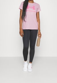 Rosa bomullst-shirt med en stor logotyp, kombinerad med svarta leggings. Vita sportskor och en beige vattenflaska kompletterar outfiten.