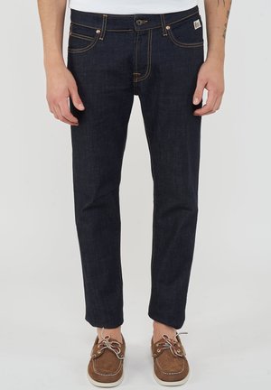 Uomo che indossa jeans blu scuro a taglio dritto e scarpe stringate in pelle scamosciata marrone, in piedi con le mani rilassate ai lati.