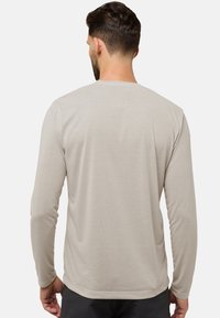 Mann mit kurzen, dunklen Haaren, der ein hellbeiges langärmeliges Hemd trägt, von hinten auf einem schlichten hellgrauen Hintergrund gezeigt.