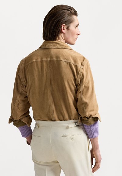 Beige Wildlederjacke mit lockerer Passform, mit hochgeschlagenen Ärmeln, die ein pastell-lila gestricktes Bündchen zeigen, kombiniert mit hellen Hose.