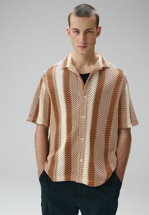 Junger Mann mit kurzen Haaren, trägt ein braun-creme gestreiftes gehäkeltes Kurzarmhemd und schwarze Hosen, die Hände in den Taschen.