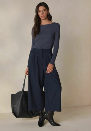 Dunkelgraues Langarmshirt kombiniert mit weitem marineblauen Hosen, schwarzen Stiefeletten bis zum Knöchel und einer großen, strukturierten schwarzen Tragetasche. Glatter Stoff und taillierte Passform.