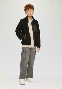 Schwarze Fleecejacke mit grauen Einsätzen, mit Reißverschluss vorne und seitlichen Taschen, getragen über einem beigen T-Shirt und kombiniert mit grauen Cargo-Hosen und weißen Sneakers.