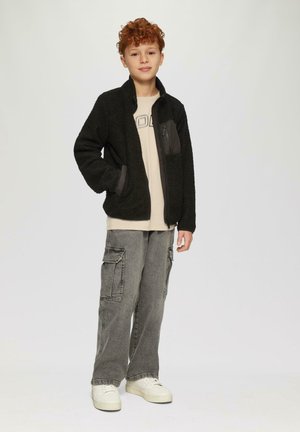 Schwarze Fleecejacke mit grauen Einsätzen, mit Reißverschluss vorne und seitlichen Taschen, getragen über einem beigen T-Shirt und kombiniert mit grauen Cargo-Hosen und weißen Sneakers.