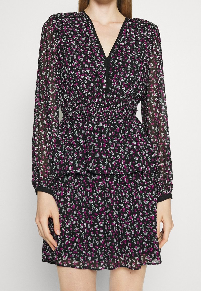 Femme portant une robe noire à manches longues avec de petits motifs floraux roses et blancs, un col en V et une taille élastique.