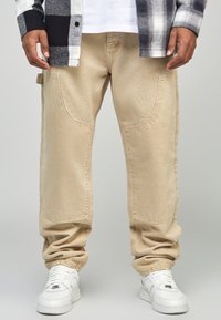 Pantaloni cargo beige con tasche laterali curve, una vestibilità rilassata e polsini a coste, abbinati a sneaker bianche e una camicia a quadri bianca e nera.